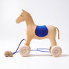 Grimm's Horse Holly is a wooden pull-along horse featuring a blue cloth saddle, four wheels, and a rope with a snail shell. Its minimalist design makes it a charming toddler toy, pictured against a plain white background.