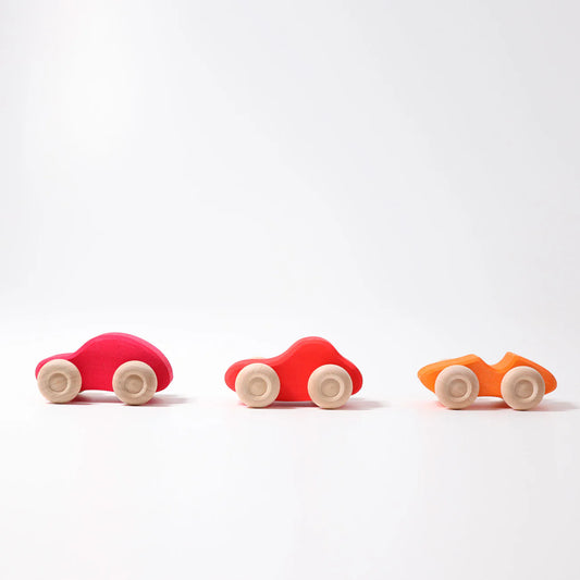 Three small Grimm's Colored Wooden Cars—pink, red, and orange—each with four light wheels, are lined up on a white surface. These non-toxic toys offer safe, colorful fun for little ones.