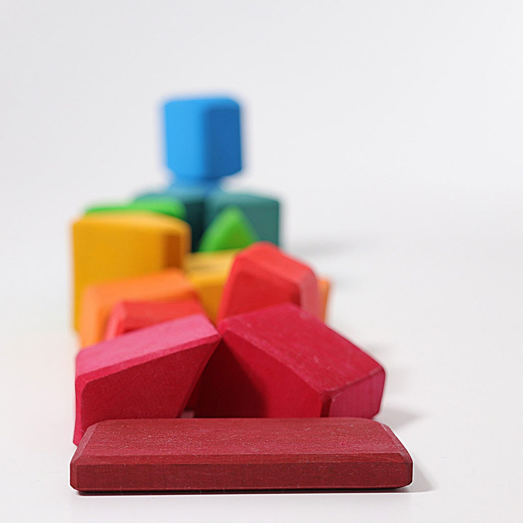 Grimm's Colored Waldorf Blocks, designed for open-ended play, feature vibrant wooden pieces arranged in a cascading pattern. These non-toxic blocks come in geometric shapes like rectangles, wedges, and cubes with red, orange, green, blue, and yellow hues against a white background.