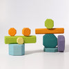 Two sets of Grimm's Tree Slices Building Blocks are stacked in playful, balanced formations on a white background. These colorful, non-toxic wooden geometric shapes, perfect for young children, include hexagons, cylinders, and elongated pieces in shades of orange, green, blue, and purple.