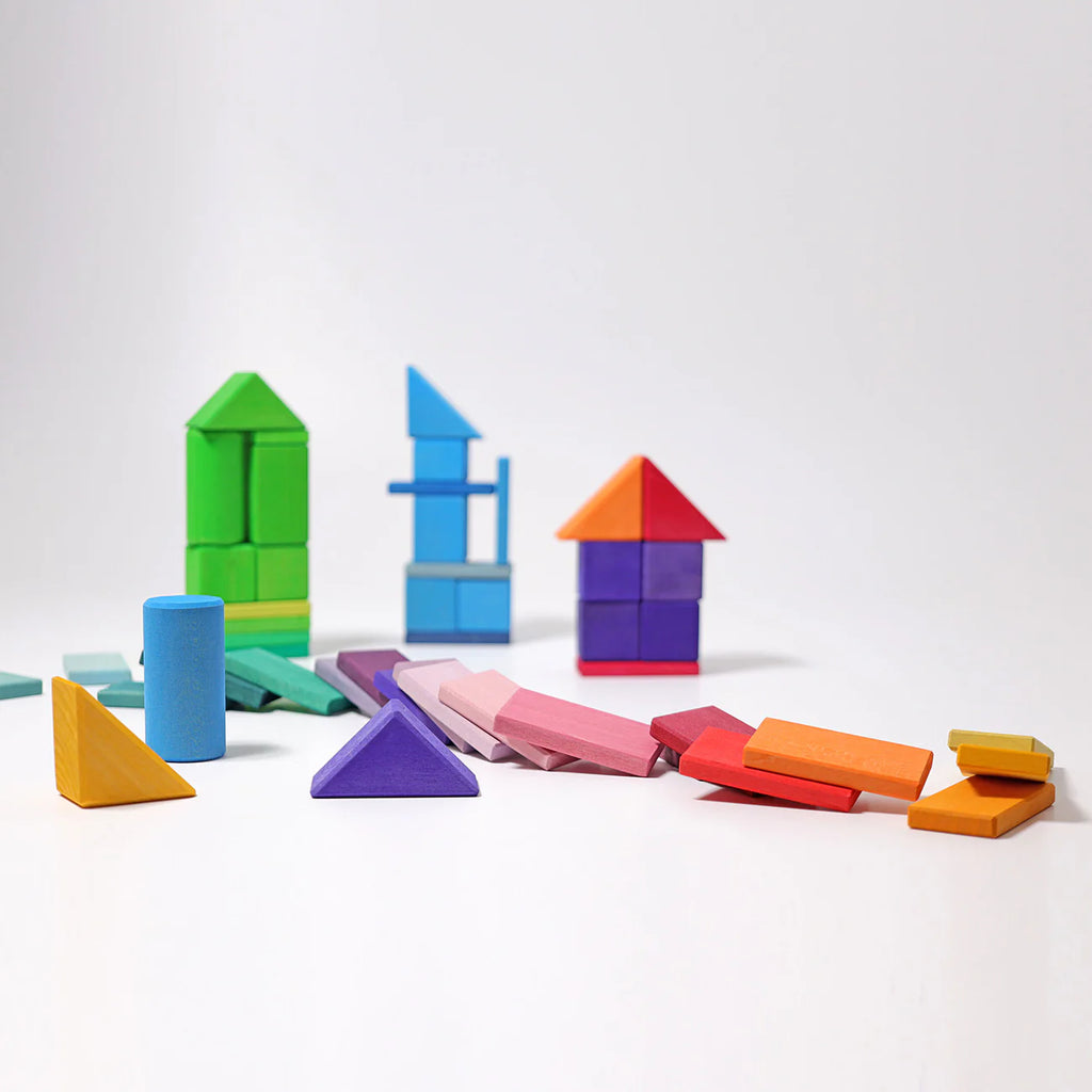Grimm's Shapes and Colors Set features vibrant geometric blocks in various shapes, arranged on a white surface—some stacked into house-like forms, others scattered—encouraging creative building and play.