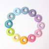 Grimm's Pastel Building Rings in various shades, reminiscent of stacking and sorting toys, arranged in a circular pattern on a white background. The rings transition through multiple colors, including blue, purple, pink, red, orange, yellow, and green, creating a rainbow-like gradient that enhances fine motor skills.