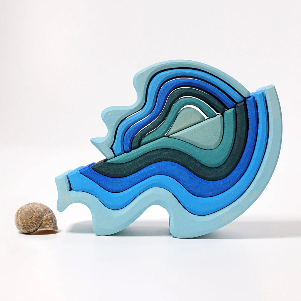 Grimm's Ocean Waves, Small—a layered wooden toy in blue and teal wave shapes—stands upright next to a small brown seashell on a plain white background.