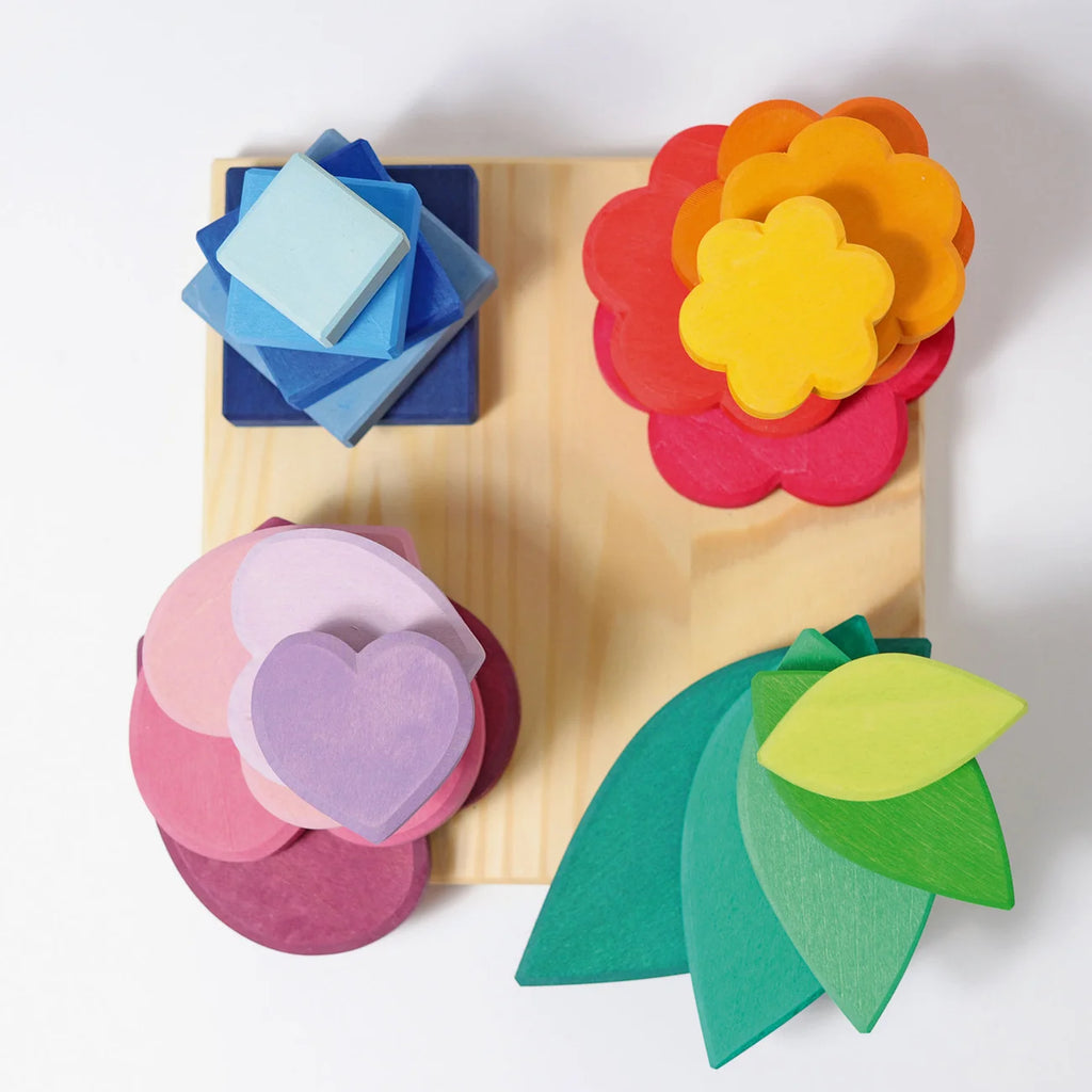 Grimm’s Stacking Game Shapes: a square wooden base featuring four sets of nesting shapes to build fine motor skills: blue squares, yellow and red flowers, pink and purple hearts, and green leaves.