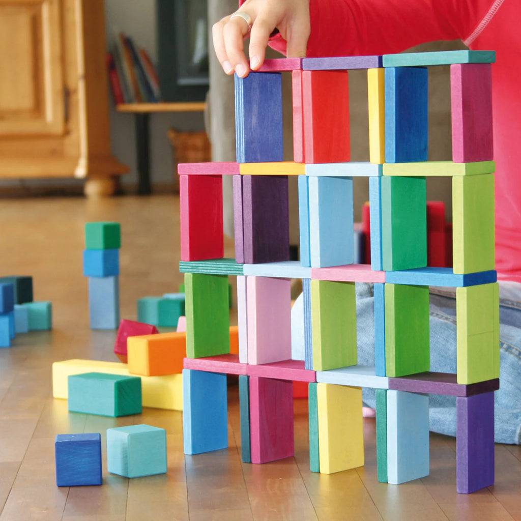 A child in a red shirt builds a tall structure on a wooden floor using Grimm's Color Charts-Rally blocks. More colorful blocks are scattered nearby, and part of the child's arm and leg are visible, demonstrating fine motor skills.