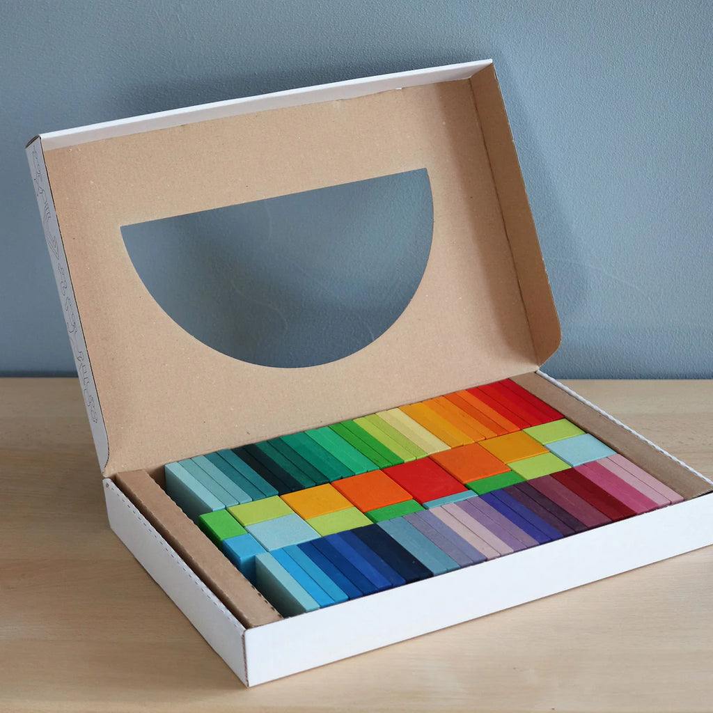 Grimm's Color Charts-Rally features an open box on a wooden surface with colorful building blocks arranged in neat, color-coordinated rows—blues, greens, yellows, reds, and purples—ideal for developing fine motor skills.