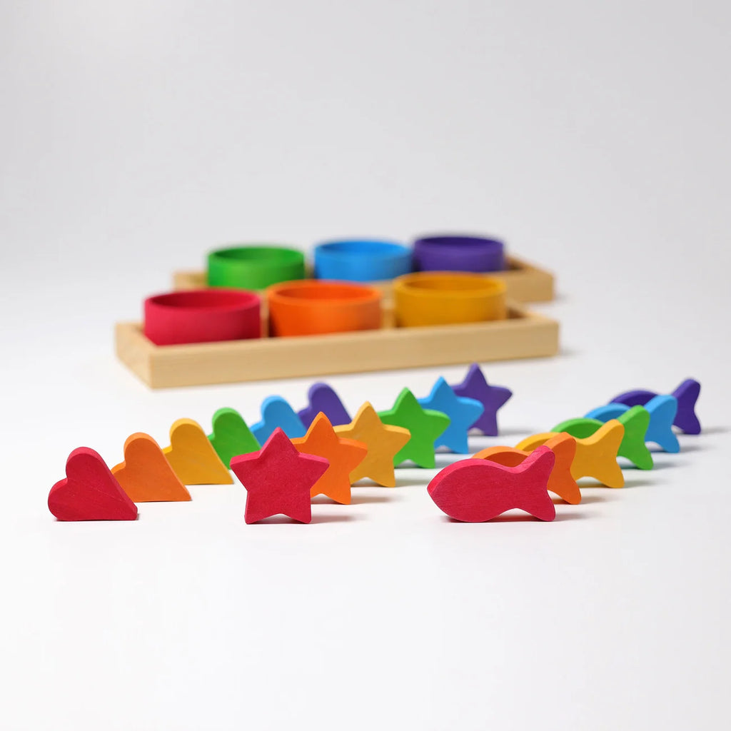 Grimm’s Sorting Game Rainbow Bowls with Tweezers featuring six colorful wooden bowls in a tray behind an assortment of rainbow-colored wooden hearts, stars, and fish used for a color sorting game.