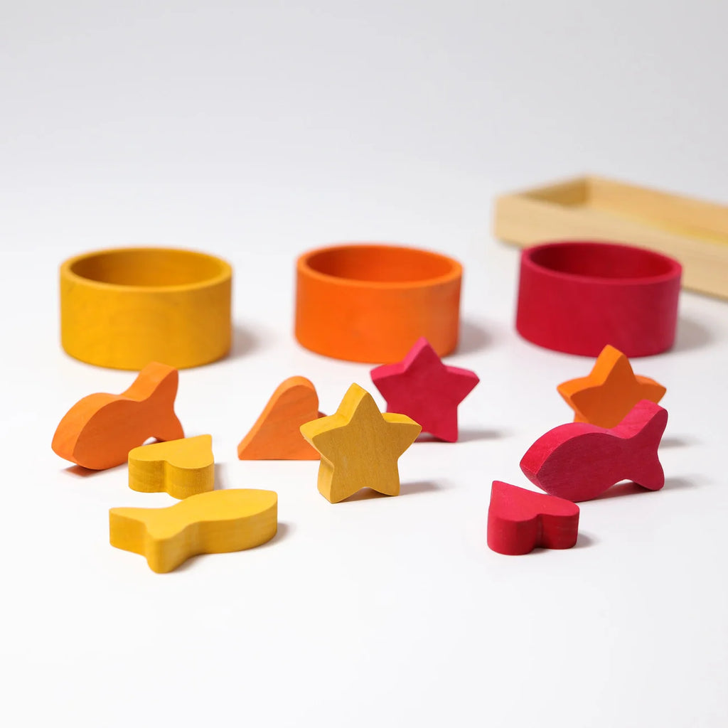Grimm’s Sorting Game Rainbow Bowls with Tweezers featuring three wooden bowls in yellow, orange, and red arranged behind a collection of matching small wooden fish, heart, and star shapes for a color sorting game.