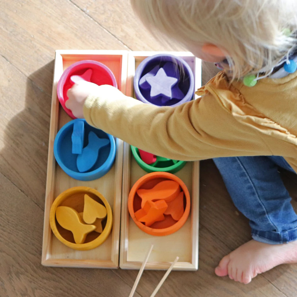 A toddler uses the wooden tweezers from a Grimm’s Sorting Game Rainbow Bowls with Tweezers set to sort wooden shapes into matching bowls, building fine motor skills.