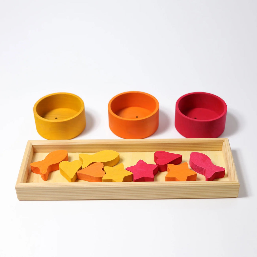 Grimm’s Sorting Game Rainbow Bowls with Tweezers featuring three wooden bowls in yellow, orange, and red arranged behind a tray of matching shapes for a rainbow sorting game.