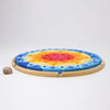 A circular wooden tray holds a colorful Grimm’s Sparkling Mandala Sun Creative Puzzle made of geometric blocks embedded with gemstone-like accents, next to a small snail shell.