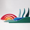 A collection of colorful, curved wooden pieces in the shapes of waves and arches, resembling landscape building blocks, are arranged artistically against a plain white background. The vibrant colors include red, orange, yellow, light blue, blue, and green. This is the Grimm's Creative Puzzle Landscape Block Set.