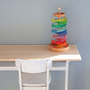 A wooden desk and white chair sit against a plain light blue wall. On the desk is the Grimm's Assembly Set Musical Ball Run, a colorful marble race set with rainbow-colored tracks and orange marbles at the base.