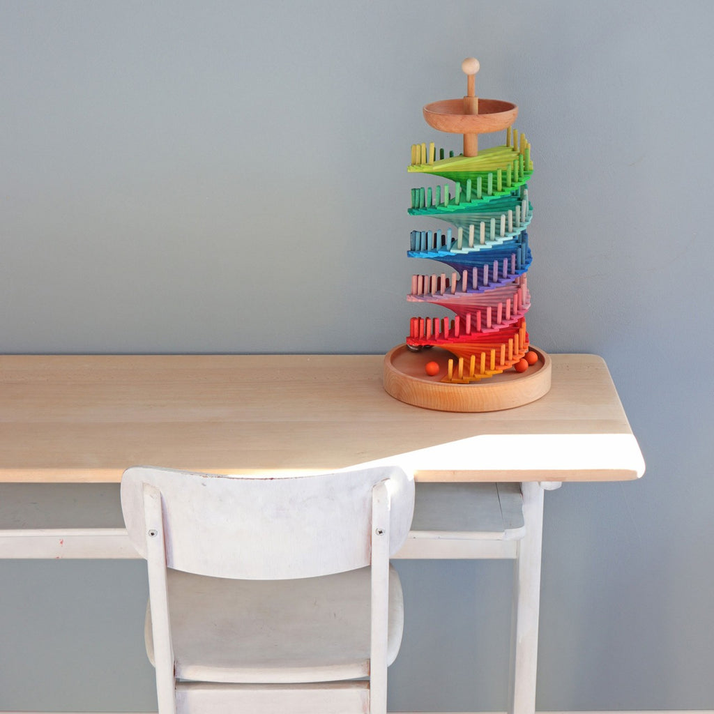 A wooden desk and white chair sit against a plain light blue wall. On the desk is the Grimm's Assembly Set Musical Ball Run, a colorful marble race set with rainbow-colored tracks and orange marbles at the base.