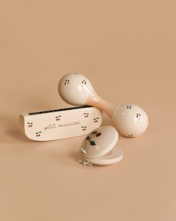 The Konges Sloejd Petit Music Set - Glossy Cherry features three cream-colored wooden instruments with cherry designs—a maraca, a harmonica case labeled "petit musicien," and a clamshell castanet—ideal for any kids’ music set.
