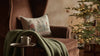 A Coral & Tusk Christmas Tree Sled Pillow, featuring embroidered animals pulling a Christmas tree on a sled, resting on a brown armchair.