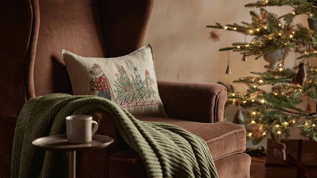 A Coral & Tusk Christmas Tree Sled Pillow, featuring embroidered animals pulling a Christmas tree on a sled, resting on a brown armchair.