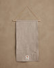 A neutral-colored linen Coral & Tusk Holiday House Advent Calendar suspended from a wooden rod with a thin cord, featuring a small patch at the bottom with an illustration of a coral branch and a fox.