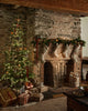 A cozy stone fireplace decorated with garland, stockings, and a Coral & Tusk Mouse with Wreath Ornament sits beside a glowing Christmas tree adorned with ornaments and presents, creating a festive holiday scene.