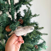 A hand holds the Finger Puppet: Shark, a small gray felt ornament, in front of a green artificial Christmas tree with pinecones—perfect for imaginative play or adding an underwater touch to your holiday decor.