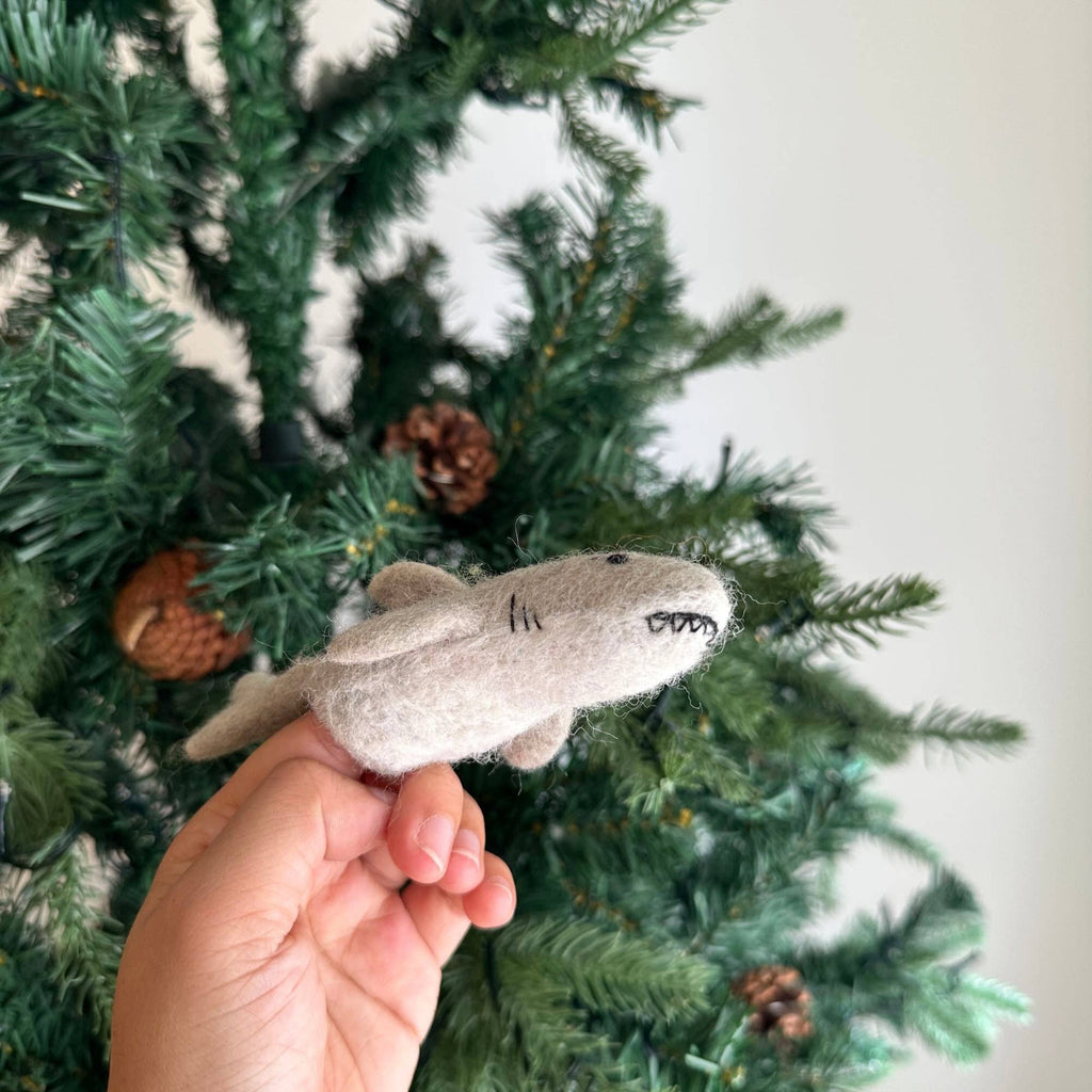 A hand holds the Finger Puppet: Shark, a small gray felt ornament, in front of a green artificial Christmas tree with pinecones—perfect for imaginative play or adding an underwater touch to your holiday decor.