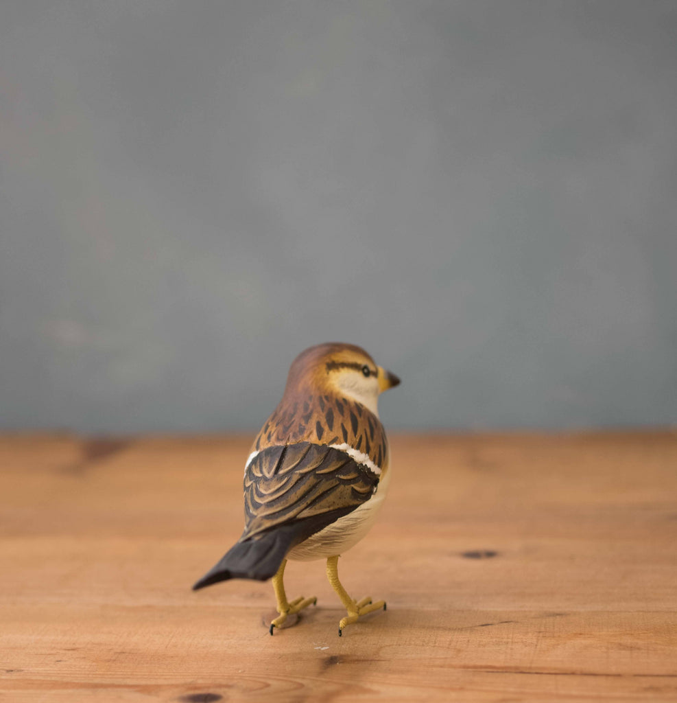The Hand Carved Ornament - Sparrow Bird - 3.5"H features brown, black, and white feathers and stands on a wooden surface with its back to the camera against a gray background.