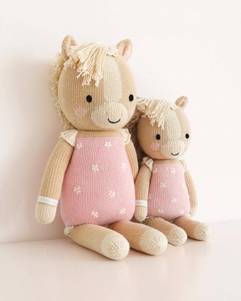Two Cuddle + Kind Sophia the Pony dolls, hand knit in Peru from fair trade materials, feature beige bodies with pink flowered outfits and manes. The ponies, one large and one small, sit side by side against a white background.