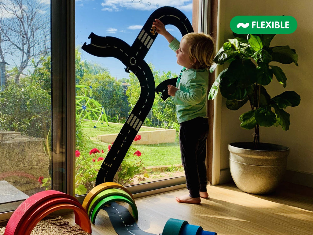A young child plays with the Large Flexible Toy Road Set - Highway (24-Piece) on a glass door. Colorful wooden arches are on the floor, a large potted plant is nearby, and a "Flexible" label appears in the upper right corner.