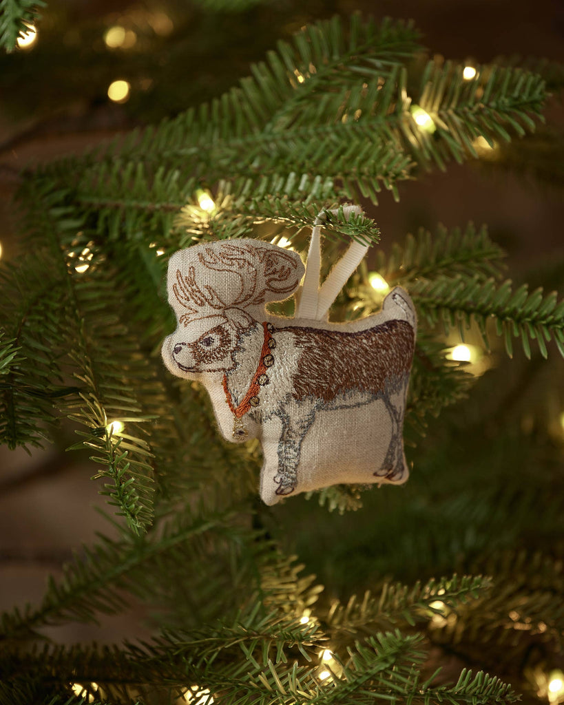 The Coral & Tusk Reindeer with Bells Ornament, featuring antlers, a red beaded necklace, and golden bells, hangs on a Christmas tree branch illuminated by warm white string lights.