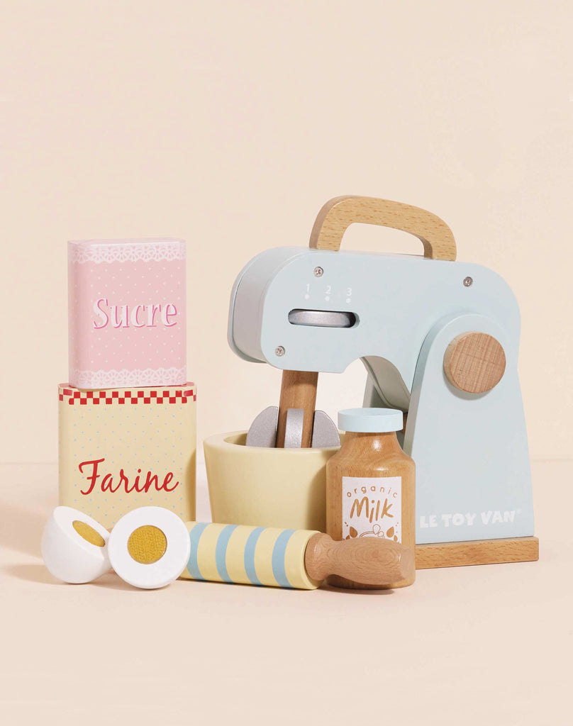The Bakers Mixer Set & Accessories is a pastel wooden toy mixer set with a mixing bowl, eggs, rolling pin, milk carton, and "Sucre" and "Farine" containers—ideal for imaginative kitchen play against a light beige background.