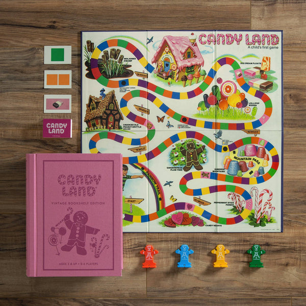 The Board Game: Candy Land Vintage Bookshelf Edition is displayed on a wooden surface, featuring its colorful board, four colored playing cards, a pink vintage bookshelf box, and gingerbread player pieces in green, yellow, blue, and orange.