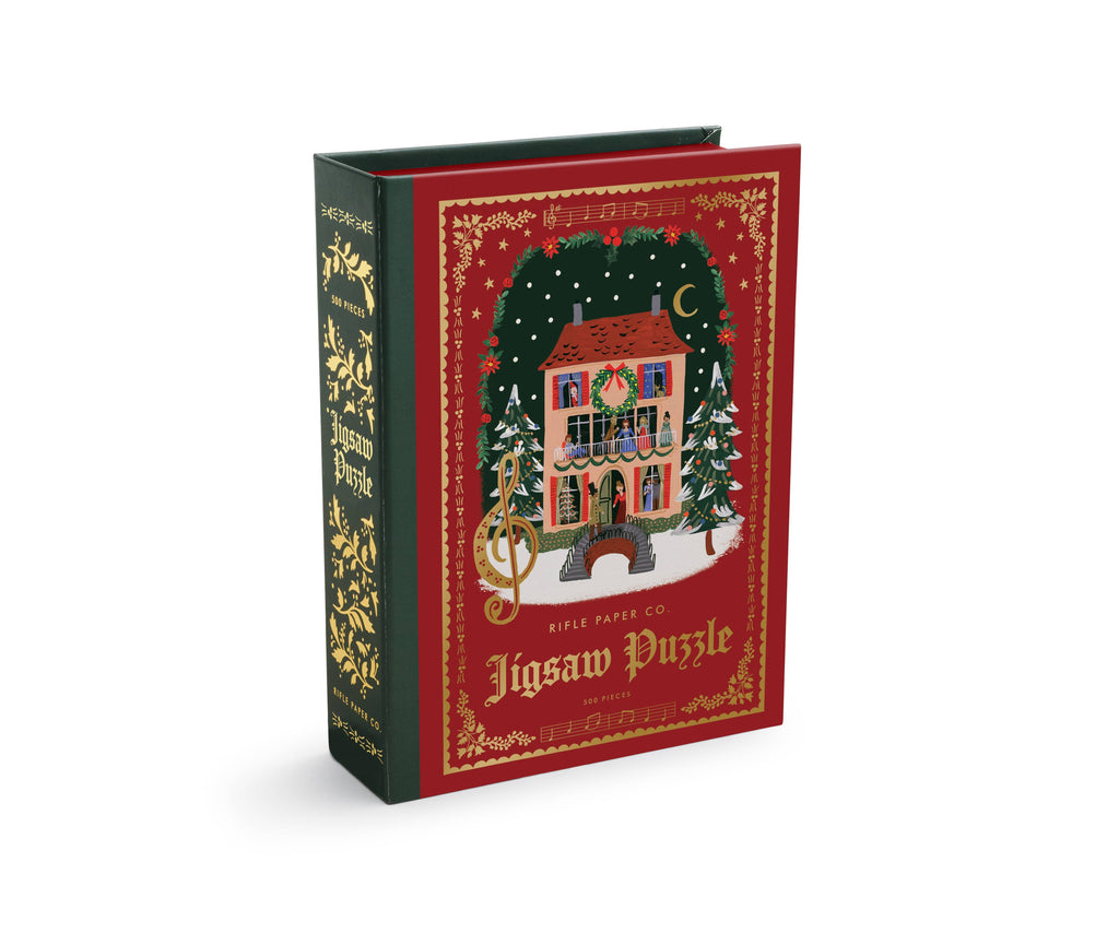 The Christmas Carols Jigsaw Puzzle is a 500-piece puzzle presented in a book-style box, featuring an illustration of a festive, snow-covered house decorated for Christmas.