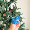 A hand holds the Finger Puppet: Whale Shark—a small, blue puppet with white spots—evoking ocean vibes before a decorated green Christmas tree with pinecones.