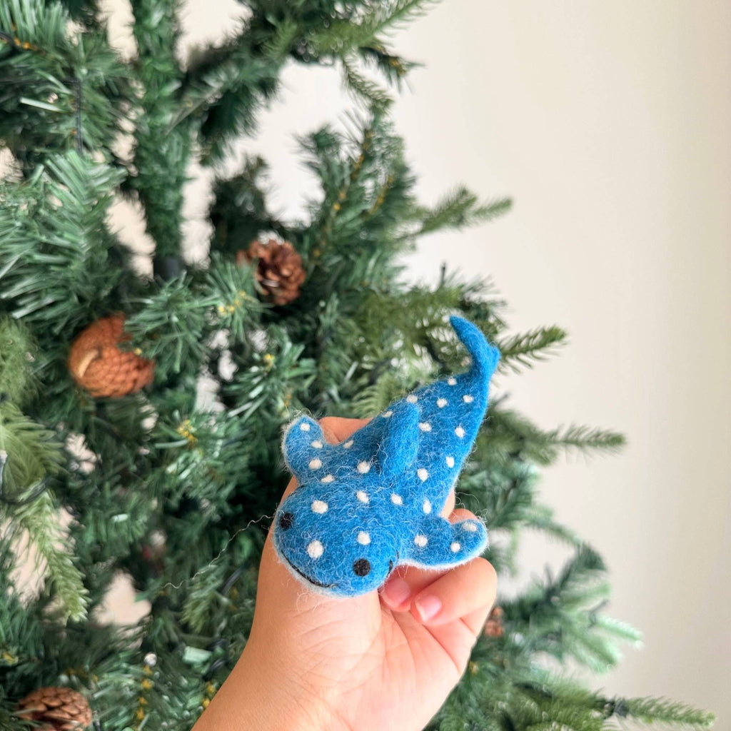 A hand holds the Finger Puppet: Whale Shark—a small, blue puppet with white spots—evoking ocean vibes before a decorated green Christmas tree with pinecones.