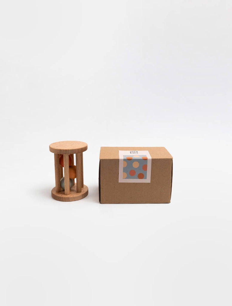The Sabo Concept Rolling Rattle - Lagoon, a small wooden baby toy with vertical bars and colored balls inside, stands beside a plain brown box labeled with blue and orange dots, both set against a white background.