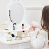 A young girl sits at a white vanity with the Vanity Star Beauty Kit, holding a pink toy hairdryer and surrounded by play makeup and accessories, her face partially reflected in the mirror.