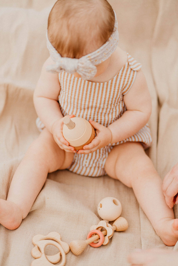 A baby in a striped romper and headband sits on a beige blanket, holding the Sabo Concept Bear Stacker - Beige. Additional wooden toys lie near the baby's legs, with the baby's face turned away from view.