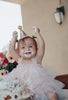 A baby wearing a pink dress and a Confetti Glitter Party Hat sits with frosting smeared on her face and hands, looking joyful with her arms raised during birthday celebrations. A partially eaten cake is in front of her, and a bouquet of flowers in the background adds charm to the photo shoot.