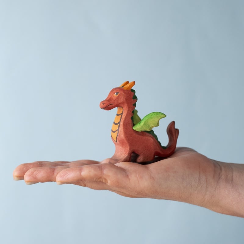 A hand holds the Bumbu Handmade Wooden Baby Dragon, a small, colorful figurine with green wings and orange details, set against a light blue background—ideal for imaginative fantasy play.