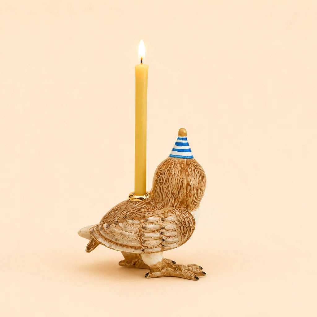 A handcrafted collectible ceramic Barn Owl Cake Topper, featuring a blue striped party hat and holding a lit yellow candle on its back, is set against a plain beige background.