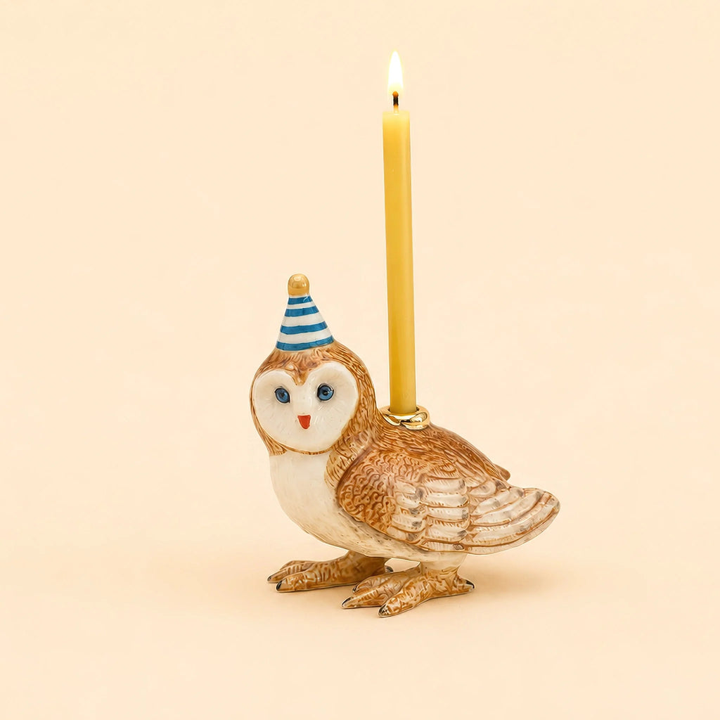 A charming Barn Owl Cake Topper, handcrafted as a collectible ceramic figurine, wears a blue and white party hat and holds a lit yellow candle on its back against a plain beige background.