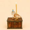 A Barn Owl Cake Topper, handcrafted from ceramic and featuring a blue striped party hat, sits on a wooden chest with a lit yellow candle on its back against a beige background.