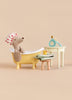 A Maileg Tricycle Mouse wearing a polka-dot shower cap sits in a miniature yellow Maileg Bathtub Set, with a small stool, towels, a rubber duck, and a toy grooming set nearby.