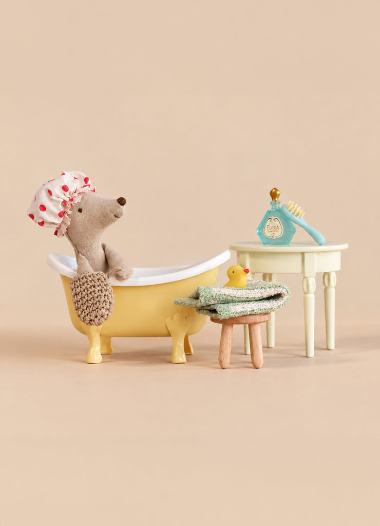 A Maileg Tricycle Mouse wearing a polka-dot shower cap sits in a miniature yellow Maileg Bathtub Set, with a small stool, towels, a rubber duck, and a toy grooming set nearby.