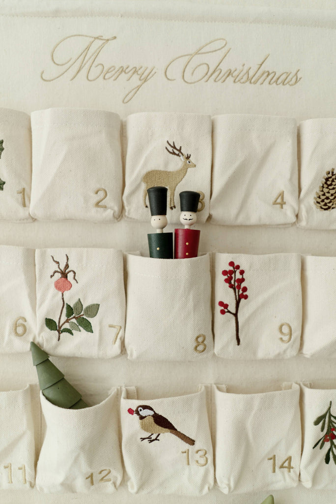 Wooden Soldiers advent calendar with embroidered pockets numbered 1 to 14, showcasing holiday motifs such as a reindeer, pinecone, and festive plants. Two wooden soldier figurines are tucked into pocket number 8.