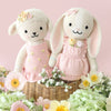 Cuddle + Kind Eleanor the Lamb, a hand-knit doll in a pink dress, sits in a wicker basket with pastel flowers and greenery against a soft pink background. Each doll is handmade in Peru with care.