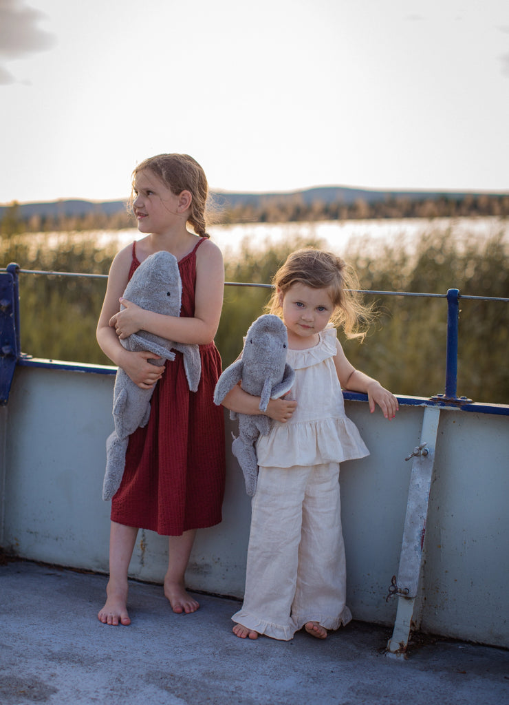 Two young girls stand on a boat deck holding a Senger Naturwelt Cuddly Animal - Shark and another plush. The older girl in red smiles off to the side, while the younger girl in white looks at the camera. Reeds and water are visible behind them.
