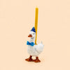 Introducing the Blue Goose Cake Topper – Final Sale: this handcrafted ceramic figurine features a goose wearing a blue bow tie and party hat, carrying a yellow candle on its back, beautifully photographed against a plain beige background.
