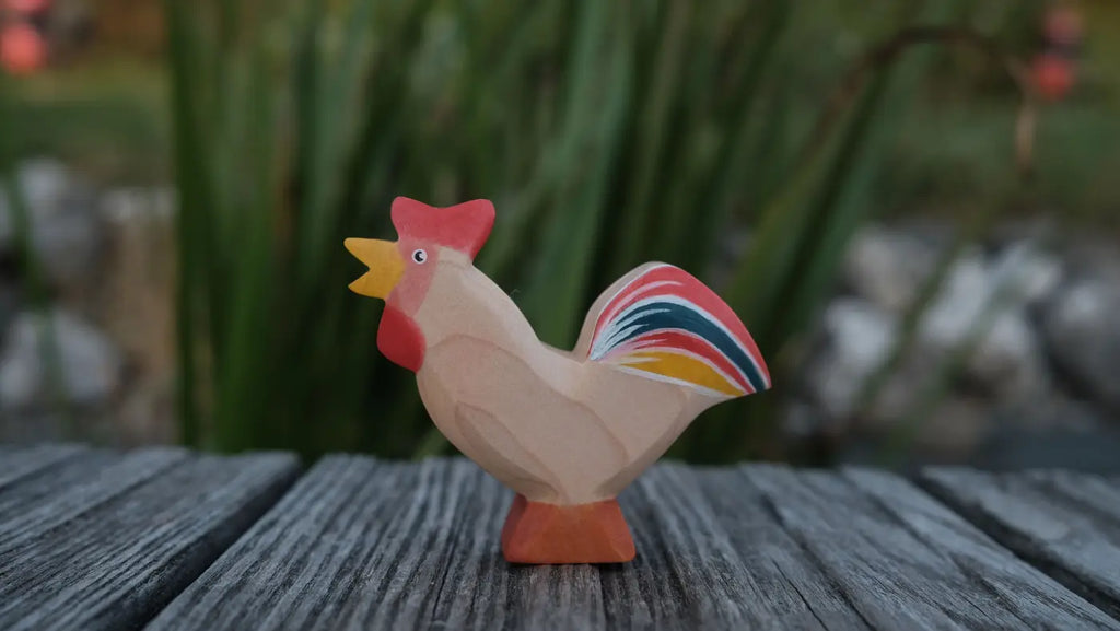 A small handmade Holzwald Bremer Rooster wooden figurine with a red comb and a multi-colored tail, standing on a wooden surface.