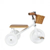 The Banwood Toddler Trike is a minimalist white tricycle with a brown seat and handlebars, featuring smooth white wheels and a front wicker basket for toddlers.